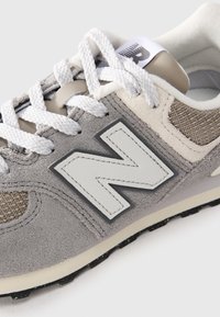 Gros plan sur une sneaker New Balance grise et beige avec des lacets blancs et un grand logo "N" brodé sur le côté.