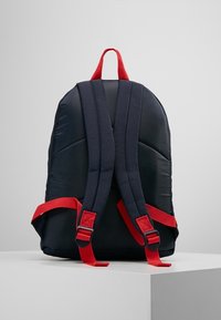 Tmavě modrý polstrovaný batoh s červenými nastavovacími popruhy a horním madlem; má hladký povrch a zaoblený design.