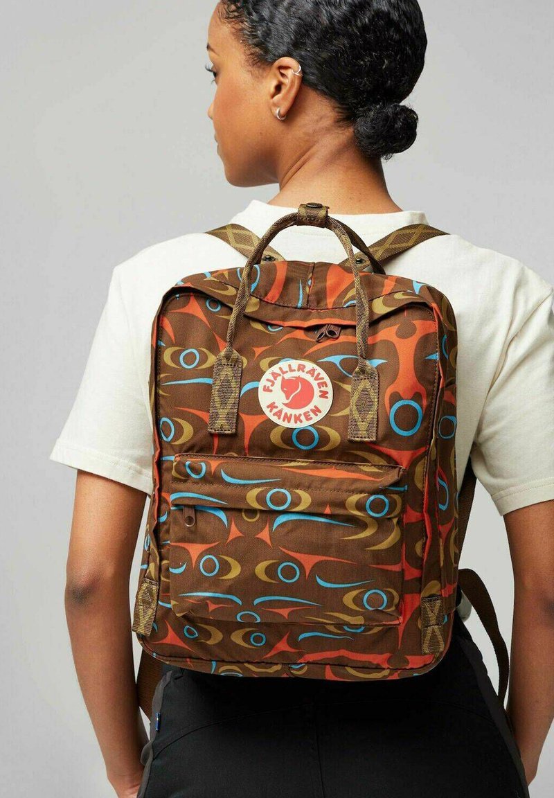 Brauner Rucksack mit gemustertem Design, der blaue und rote abstrakte Motive zeigt. Er verfügt über eine vordere Reißverschlusstasche, einen oberen Griff und verstellbare Schultergurte.
