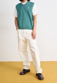 Persoon in een teal mouwloos vest over een wit T-shirt, crèmekleurige broek en donkerpaarse schoenen, staand op een mosterdkleurige vloer.