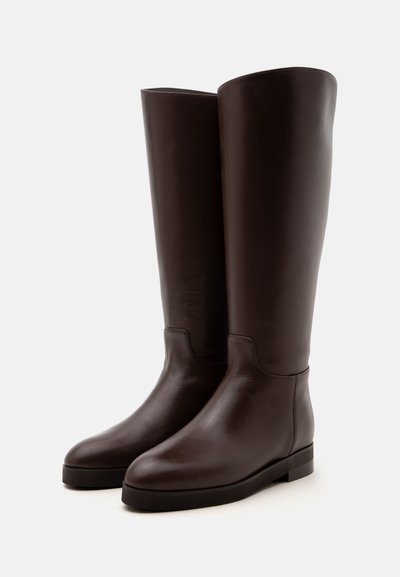 Paire de bottes hautes en cuir marron foncé avec des bouts arrondis et des semelles plates sur un fond clair uni.