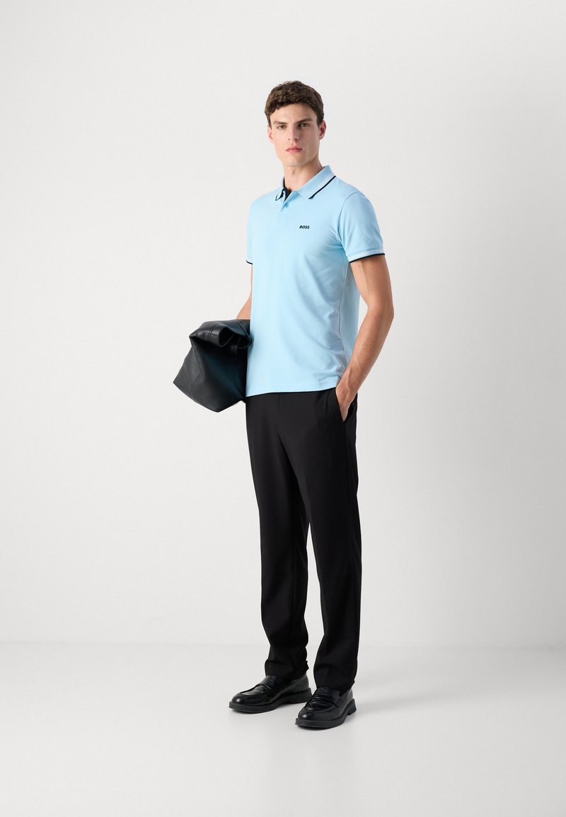 Polo bleu clair avec des bordures noires, logo sur la poitrine, associé à un pantalon noir et des chaussures noires, tenant un sac noir.