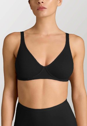 Donna che indossa un semplice reggiseno nero senza ferretto con spalline sottili, mostrata dalla metà del petto al collo su uno sfondo chiaro uniforme.
