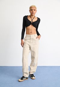 Zwarte lange mouwen top met draai-detail en beige wijde broek; model draagt zwarte en crèmekleurige sneakers op een lichtblauwe vloer.