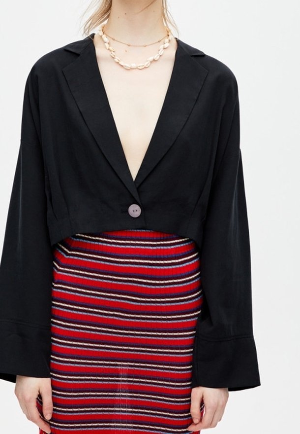 Černá zkrácená bunda s výstřihem do V a širokými rukávy, zapínaná fialovým knotem. Kombinována s barevnými pruhovanými sukněmi v červené a modré.
