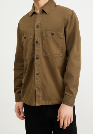 Homme portant une chemise marron à boutons avec deux poches poitrine, dont une brodée du mot BOSS, associée à un pantalon noir.