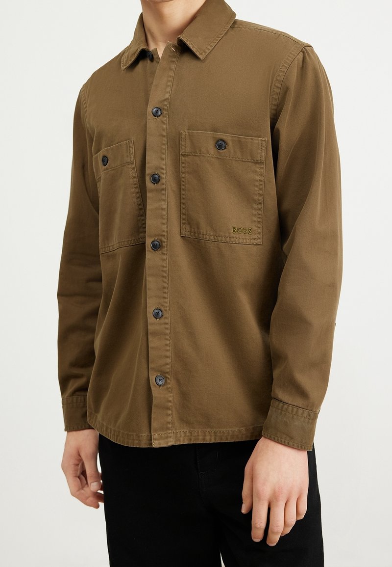 Homme portant une chemise marron à boutons avec deux poches poitrine, dont une brodée du mot BOSS, associée à un pantalon noir.