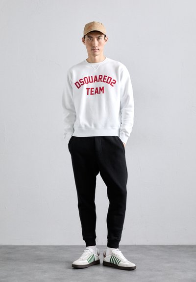 Felpa bianca con testo rosso "DSQUARED2 TEAM", pantaloni neri, e sneakers bianche con strisce verdi e suole marroni. Cappello beige in testa.