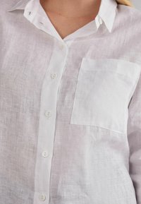 Bílá lněná košile na knoflíky s kapsou na prsou, s mírně texturovaným povrchem a klasickým límcem, zapínaná na bílé knoflíky.