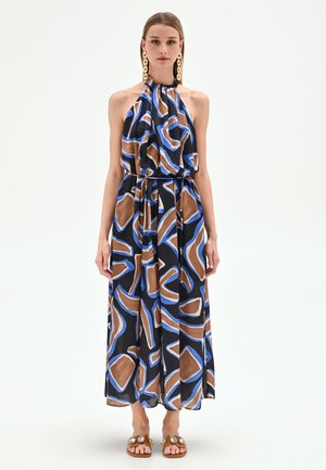 Femme portant une robe maxi dos nu sans manches avec un motif géométrique abstrait bleu, marron et beige, associée à des boucles d'oreilles dorées imposantes et des sandales plates.