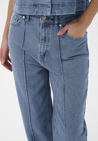 Frau in hellblauen Jeans mit sichtbaren Nähten, eine Hand in der Tasche, und einem passenden, kurz geschnittenen Denim-Oberteil vor einem schlichten Hintergrund.