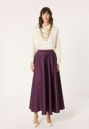 Žena nosi krémovú blúzku, vrstvené zlaté náhrdelníky, vysoký pás fialovej maxi sukne a krémové lodičky so špicatou špičkou na bielom pozadí.