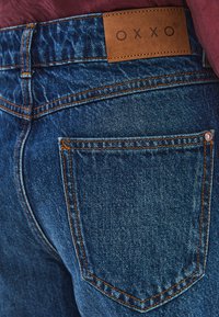 Denimjeans i en medelblå nyans med orange sömmar. Har en brun läderlogga på midjan och två bakfickor.