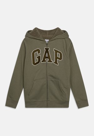 Bluza z kapturem na zamek w kolorze oliwkowej zieleni, z ociepleniem z polaru, przednimi kieszeniami oraz dużymi literami „GAP” na klatce piersiowej w ciemnobrązowym kolorze z białym obrysem.