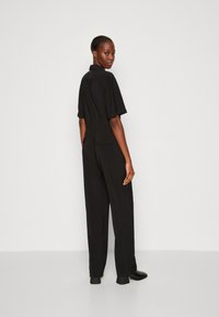 Jumpsuit noir à manches courtes, coupe décontractée, couture à la taille et poches au dos. Texture lisse et design à jambes larges ; porté avec des chaussures noires.