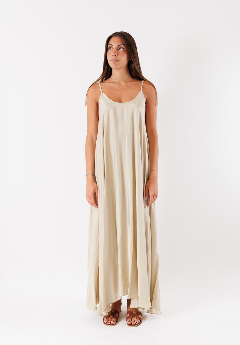 Abito maxi beige con spalline sottili, silhouette fluida, tessuto morbido e orlo asimmetrico, abbinato a sandali marroni.