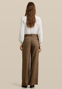 Blouse blanche avec des détails de manches froncées, associée à un pantalon large en tweed marron taille haute, avec des poches arrière et une étiquette au niveau de la taille.