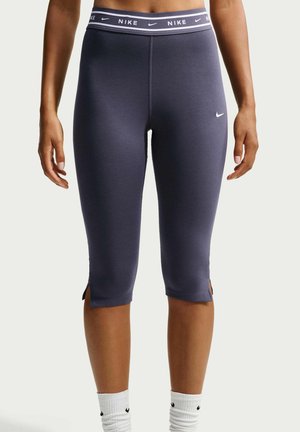 Vrouw draagt marineblauwe Nike cropped leggings met gespleten zomen, Nike-logo op linker dij en een gebrande elastische tailleband.