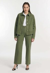 Olijfgroen denimjack met twee voorkruiszakken, zilveren drukknopen en bijpassende wijde broek. Witte t-shirt eronder. Zwarte hoge hakken.