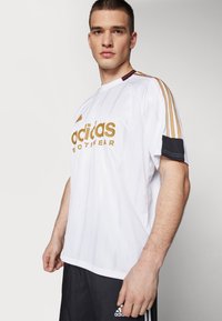 Balta Adidas sportinė marškinė su auksiniu emblemą, vertikaliomis juostomis ir juodu apvadu ant rankovių; dėvima su juodomis šortais.