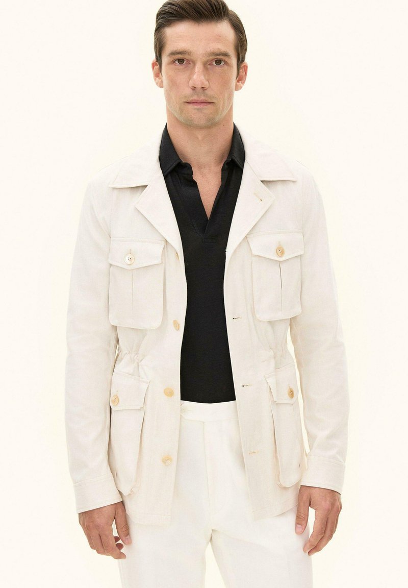 Uomo che indossa una giacca safari beige con quattro tasche frontali e una camicia nera con collo, su uno sfondo semplice.