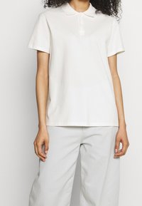 Polo shirt in light beige cotton with a classic collar, short sleeves, and a subtle logo accent on the chest. Paired with light gray pants.