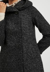 Kofta i ullblandning i strukturerat svart och grått med huva, enkel knappstängning och vida ärmar. Har ett prickigt stickad mönster.