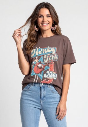 Donna sorridente con capelli castani ondulati che indossa grandi orecchini a cerchio colorati, una maglietta marrone con stampa grafica e jeans azzurri, posando davanti a uno sfondo semplice.