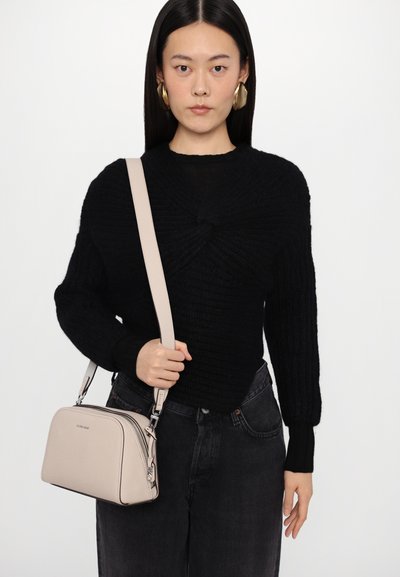 Black knitted sweater with a twisted design, paired with high-waisted black jeans and a light beige structured handbag. Fashionable gold earrings.