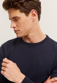 Junger Mann mit kurzen braunen Haaren, der einen dunkelblauen Rundhalsausschnitt-Pullover trägt, nach unten schaut und seinen Ärmel auf einem neutralen Hintergrund anpasst.