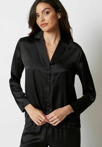Zwarte zijden pyjamakey met lange mouwen, knoopsluiting en een klassieke kraag. De gladde textuur en subtiele glans versterken de elegante uitstraling.