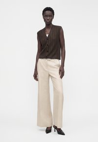 Femme portant un gilet sans manches marron foncé boutonné, un pantalon beige à jambes larges et des talons noirs à bout pointu, debout sur un fond blanc.