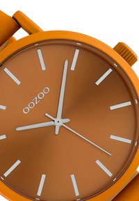 Orange silicone wristwatch featuring a matte round face, silver hour markers, and hands, with a prominent logo positioned at the center.