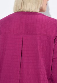 Fuksia takki, jossa on teksturoitu, ryppyinen muotoilu; siinä on takasauma, kauluskäänne ja sileä pinta. Näkyy takaa.