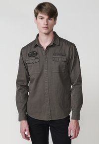 Donkergrijs overhemd met lange mouwen en knoopsluiting, met twee borstzakken en geborduurde patches. Glad materiaal, klassieke pasvorm.