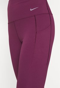 Zbliżenie na fioletowe legginsy z wysokim stanem, z boczną kieszonką i odblaskowym, srebrnym logo Nike na pasku.