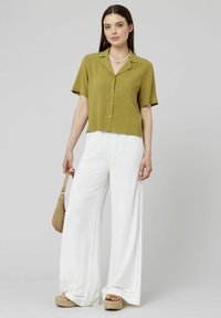 Chemise à manches courtes olive encolure ouverte, associée à un pantalon large blanc avec des piqûres sur le côté. Accessoire en sac tissé.