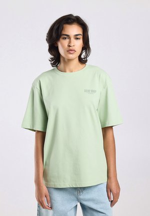 Persoon met schouderlang haar, die een lichtgroen oversized T-shirt met de tekst "SURF SHOP" en lichtblauwe spijkerbroek draagt, staand tegen een effen achtergrond.