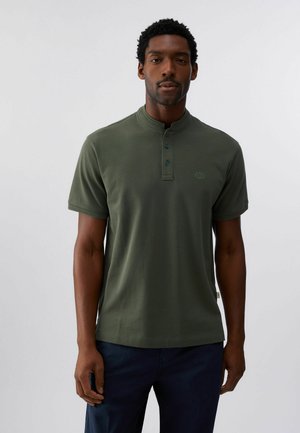 Homme portant un t-shirt henley manches courtes vert foncé avec trois boutons, associé à un pantalon bleu foncé, se tenant devant un fond uni.