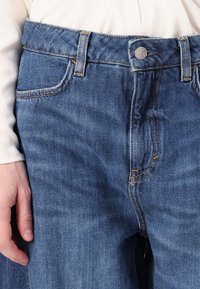 Niebieskie dżinsy mają klasyczny pięciopocketowy fason z żółtym przeszyciem, srebrnym zapięciem na guzik oraz luźnym krojem w okolicy talii.