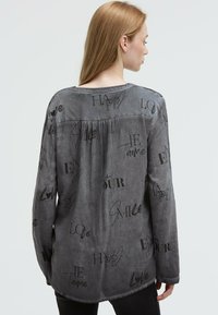 Frau mit langen blonden Haaren, die ein graues Langarmshirt trägt, bedruckt mit schwarzen Wörtern wie "Liebe", "Glücklich" und "Je t’aime."