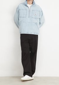 Ljusblå denimpullover med halvdragkedja, två framfickor och en logotypapplikation. Matcher med svarta löst sittande byxor och vita skor.