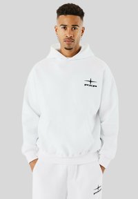 Jonge man met sproeten die een witte hoodie en bijpassende trainingsbroek draagt, beide voorzien van een klein zwart "PXP" logo, met de handen in zijn zakken.