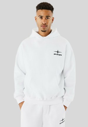Giovane con lentiggini che indossa un felpa bianca con cappuccio e pantaloni della tuta abbinati, entrambi con piccolo logo nero "PXP", mani nelle tasche.