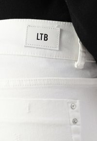 Hvit denimstoff med et lærmerke som leser "LTB". Inkluderer sølvfarget knappemateriale og sømmer. Nærbilde.