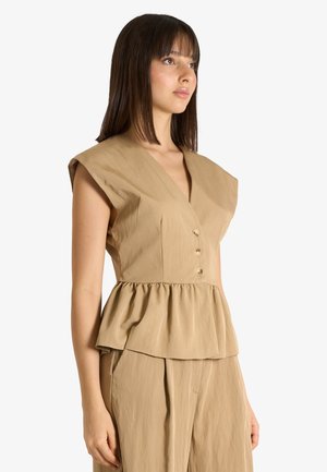 Donna con capelli castani lisci che indossa un top beige senza maniche con bottoni e vita peplum abbinato a pantaloni ampi coordinati.