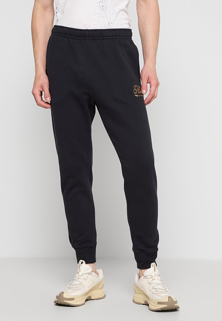 Schwarze Sweatpants mit elastischem Bund, schmal zulaufenden Beinen und goldener Stickerei auf der linken Seite. Hergestellt aus weichem Baumwollmaterial.