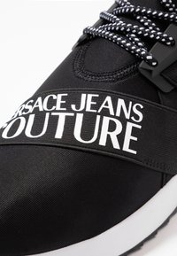 Svart idrottssko med en texturerad ovandel, vit gummisula och en framträdande vit logoband över mellanfoten, samt svarta och vita snören.