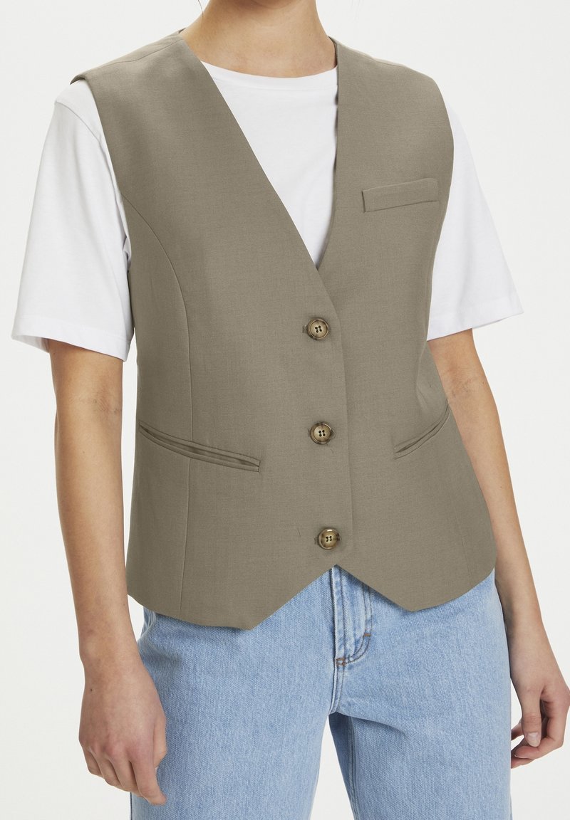 Beige väst med djup V-ringning, fyra knappar och en liten bröstficka. Bärs över en vit t-shirt, matchad med ljusblå jeans.