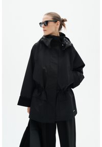 Femme aux cheveux tirés en arrière, portant une veste à capuche noire oversize, un pantalon noir et des lunettes de soleil foncées sur fond blanc uni.